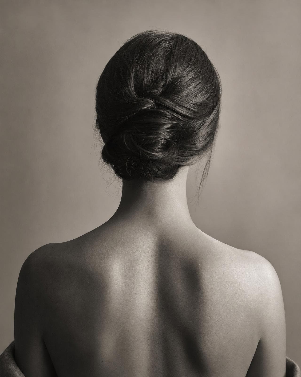The nape and bare shoulders of a woman with her hair in a sculptural updo, soft light tracing gentle curves and clean lines
