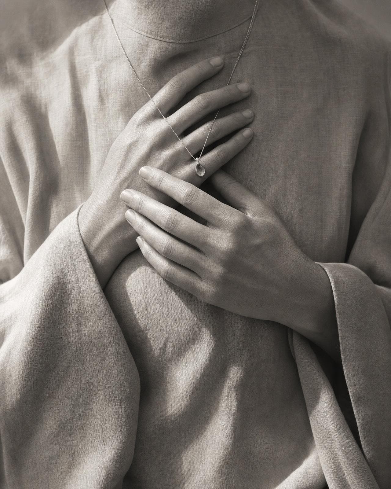 Two hands pressed gently over the heart against draped fabric, a delicate pendant chain glinting between the fingers