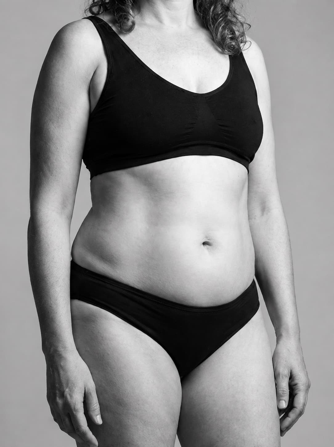 Black-and-white photograph of a woman's midsection in a plain black bra and briefs, standing with her arms relaxed at her sides