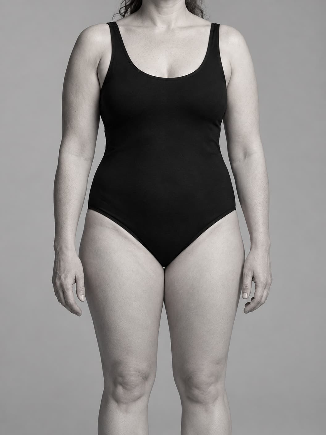 Monochrome study of a woman in a black swimsuit with her upper arms and thighs visible against a neutral gray background