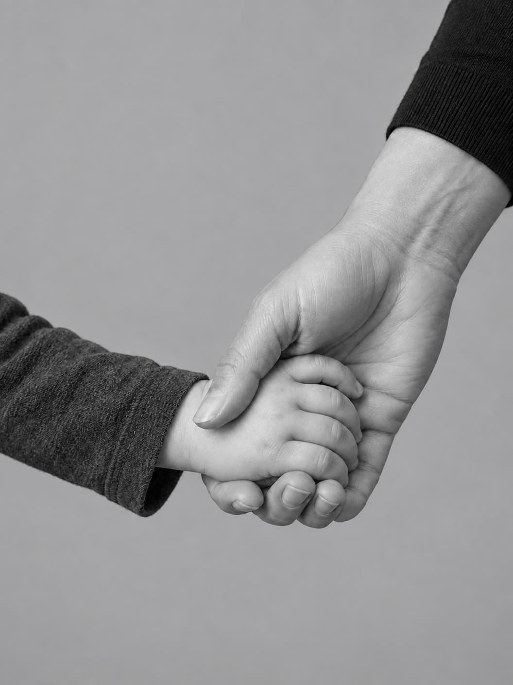 Monochrome close-up of a parent’s hand gently supporting a child’s hand against a neutral gray background